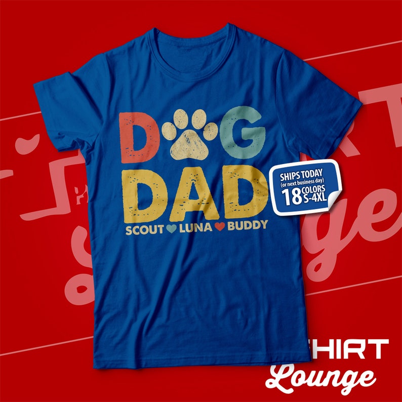 May include: Blue t-shirt with a vintage-style graphic that says "Dog Dad" with a paw print in the "O". The graphic is in red, yellow, blue, and green. Below the graphic, it says "Scout Luna Buddy" with a heart between each name.