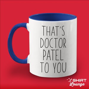Pode incluir: Caneca de cer&acirc;mica branca com borda e al&ccedil;a azuis. A caneca tem texto preto que diz "That's Doctor Patel to you".
