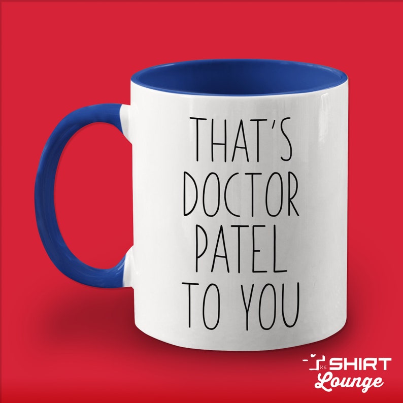 Pode incluir: Caneca de cer&acirc;mica branca com borda e al&ccedil;a azuis. A caneca tem texto preto que diz "That's Doctor Patel to you".