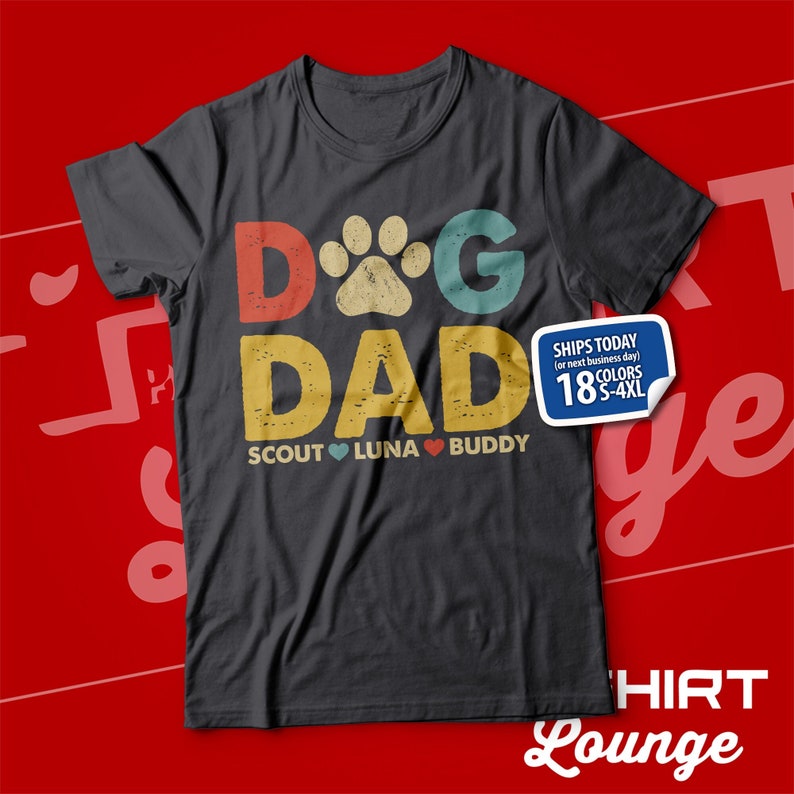 May include: A dark grey t-shirt with a vintage-style graphic that says "Dog Dad" with a paw print in the "O". The graphic is in red, yellow, blue, and green. Below the graphic, it says "Scout Luna Buddy" with a heart between each name.