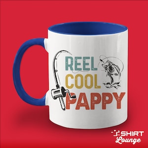 May include: White ceramic coffee mug with a blue handle and rim. The mug features a vintage-style graphic of a fishing rod, a fish, and the text "REEL COOL PAPPY".