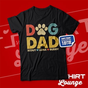 May include: Black t-shirt with a vintage-style graphic that says "Dog Dad" with a paw print in the "O". The graphic is in red, yellow, and blue. Below the graphic, it says "Scout Luna Buddy" with a red heart between each name.