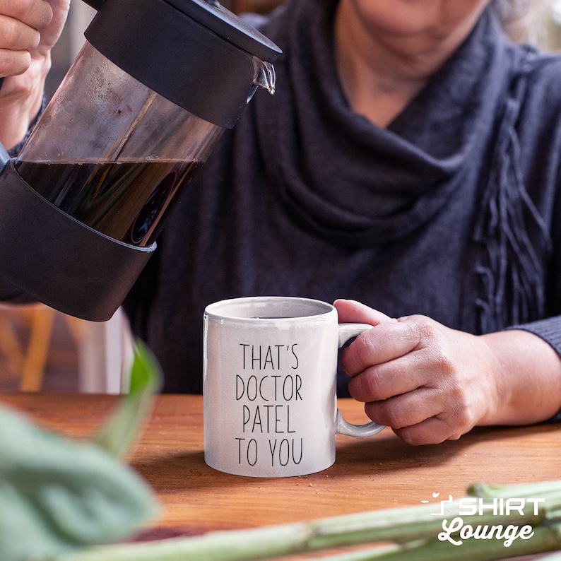 Pode incluir: Caneca de caf&eacute; de cer&acirc;mica branca com texto preto que diz "That's Doctor Patel to you". Uma pessoa est&aacute; despejando caf&eacute; de uma prensa francesa na caneca.