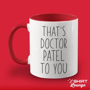 Pode incluir: Caneca de cer&acirc;mica branca com borda e al&ccedil;a vermelhas. A caneca tem texto preto que diz "That's Doctor Patel to you".
