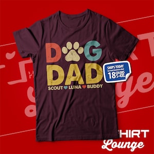 May include: A maroon t-shirt with a vintage-style graphic that says "Dog Dad" with a paw print in the "O". The graphic is in shades of blue, green, red, and yellow. Below the graphic, it says "Scout Luna Buddy" with a heart between each name.