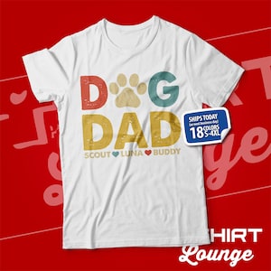 May include: White t-shirt with a vintage-style graphic that says "Dog Dad" with a paw print in place of the "O". The text is in red, blue, and green. Below the text, it says "Scout Luna Buddy" with hearts between the names.