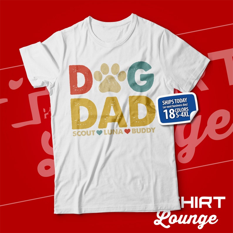 May include: White t-shirt with a vintage-style graphic that says "Dog Dad" with a paw print in place of the "O". The text is in red, blue, and green. Below the text, it says "Scout Luna Buddy" with hearts between the names.