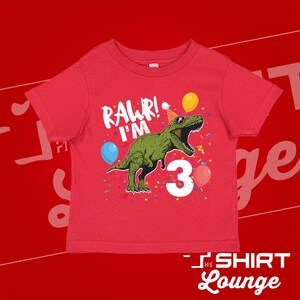 May include: Red short-sleeved t-shirt with a green dinosaur wearing a party hat and sunglasses. The dinosaur is surrounded by confetti and balloons. The text "RAWR! I'M 3" is printed on the shirt.
