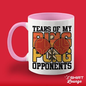May include: White ceramic coffee mug with a pink handle. The mug has a vintage-style graphic of a ping pong paddle and ball with the text "Tears of my Ping Pong Opponents".