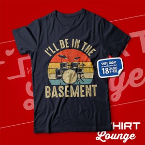 May include: A dark blue t-shirt featuring a vintage-style graphic of a drum kit in front of a sunset. The text "I'll be in the basement" is printed below the drum kit.