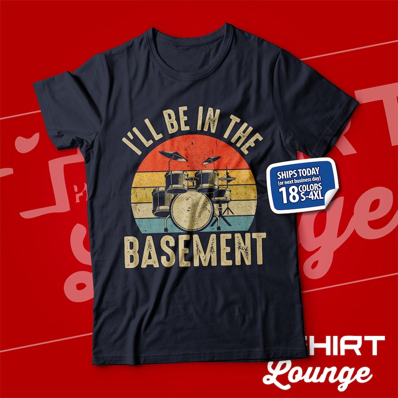 May include: A dark blue t-shirt featuring a vintage-style graphic of a drum kit in front of a sunset. The text "I'll be in the basement" is printed below the drum kit.