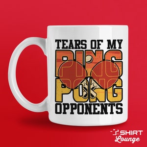 May include: White ceramic coffee mug with a graphic design of a ping pong paddle and ball. The text "Tears of my ping pong opponents" is printed on the mug.