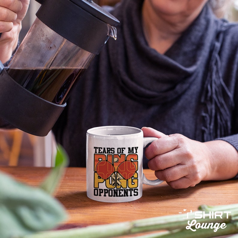 May include: White ceramic coffee mug with the text "Tears of my ping pong opponents" in orange and black lettering. The mug is filled with a brown liquid and is being held by a hand.