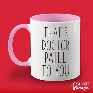 Pode incluir: Caneca de cer&acirc;mica branca com borda e al&ccedil;a rosa. A caneca tem texto preto que diz "That's Doctor Patel to you".