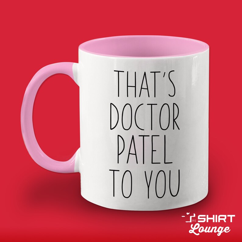 Pode incluir: Caneca de cer&acirc;mica branca com borda e al&ccedil;a rosa. A caneca tem texto preto que diz "That's Doctor Patel to you".