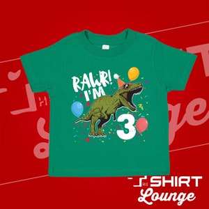 May include: Green t-shirt with a green dinosaur wearing a party hat and sunglasses. The dinosaur is holding a number 3 and there are balloons and confetti around it. The text "Rawr! I'm 3" is printed on the shirt.
