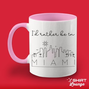 May include: White ceramic mug with a pink rim and handle. The mug has a black outline of a Miami skyline with the text "I'd rather be in Miami".