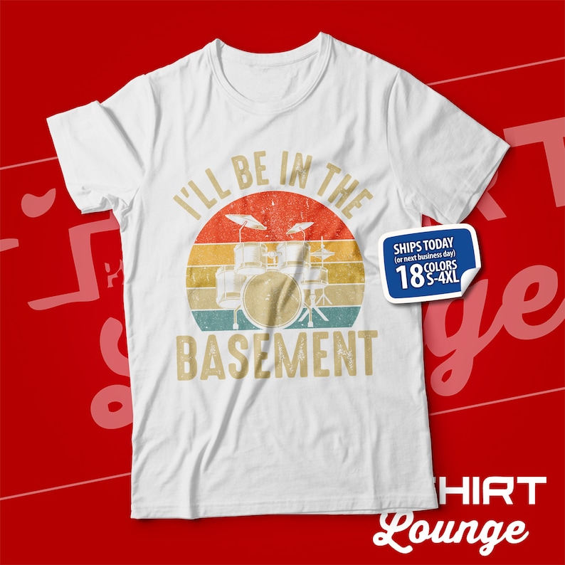 May include: White t-shirt featuring a vintage-style graphic of a drum kit in front of a sunset. The text "I'll be in the basement" is printed below the drum kit.