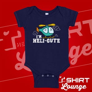 May include: A navy blue short-sleeved baby bodysuit featuring a graphic illustration of a teal helicopter with yellow rotors. Below the helicopter, the text reads "I'M HELI-CUTE" in white capital letters. The design includes small white cloud outlines.