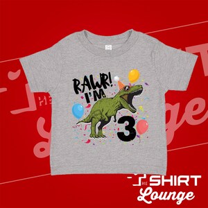 May include: Gray short-sleeved t-shirt with a green dinosaur wearing a party hat and sunglasses. The dinosaur is surrounded by confetti and balloons. The text "Rawr! I'm 3" is printed on the shirt.