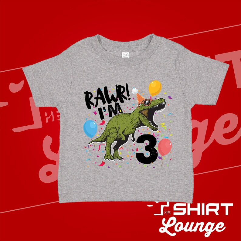 May include: Gray short-sleeved t-shirt with a green dinosaur wearing a party hat and sunglasses. The dinosaur is surrounded by confetti and balloons. The text "Rawr! I'm 3" is printed on the shirt.