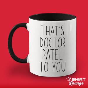 Pode incluir: Caneca de cer&acirc;mica branca com al&ccedil;a e borda pretas. A caneca tem texto preto que diz "That's Doctor Patel to you".