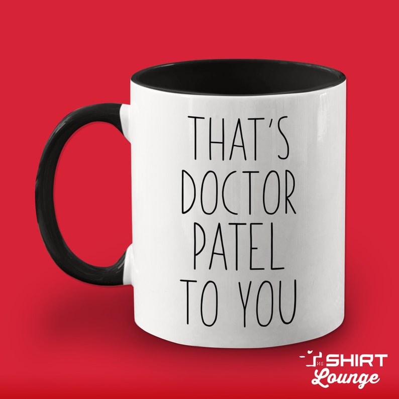 Pode incluir: Caneca de cer&acirc;mica branca com al&ccedil;a e borda pretas. A caneca tem texto preto que diz "That's Doctor Patel to you".