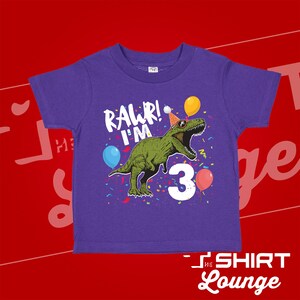 May include: Purple short-sleeved t-shirt with a green dinosaur wearing a party hat and sunglasses. The dinosaur is surrounded by confetti and balloons. The text "RAWR! I'M 3" is printed on the shirt.
