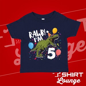 May include: A navy blue t-shirt with a green dinosaur wearing a party hat and sunglasses. The dinosaur is surrounded by confetti and balloons. The text "RAWR! I'M 5" is printed on the shirt.