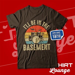 May include: A brown t-shirt featuring a vintage-style graphic of a drum kit set against a sunset backdrop. The text "I'll be in the basement" is printed beneath the drum kit. The text "Ships Today (or next business day) 18 Colours S-4XL" is printed in a white box on the right side of the shirt.