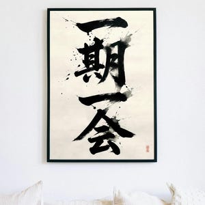 Peut inclure: Œuvre d'art encadrée avec calligraphie japonaise. Des coups de pinceau noirs audacieux se détachent sur un fond crème. La calligraphie, "Ichigo Ichie", est agrémentée d'éclaboussures d'encre. Un sceau rouge est visible en bas à droite.