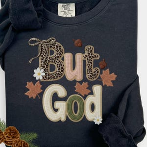 Pu&ograve; includere: Felpa nera con la frase "But God". La parola "But" presenta un motivo leopardato, mentre "God" &egrave; in verde e crema. Il design include elementi a tema autunnale come foglie e ghiande.