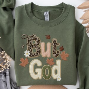 Pu&ograve; includere: Felpa verde oliva con la frase ricamata "But God". La parola "But" presenta un motivo leopardato, con accenti floreali e di foglie. La felpa ha un girocollo e maniche lunghe.