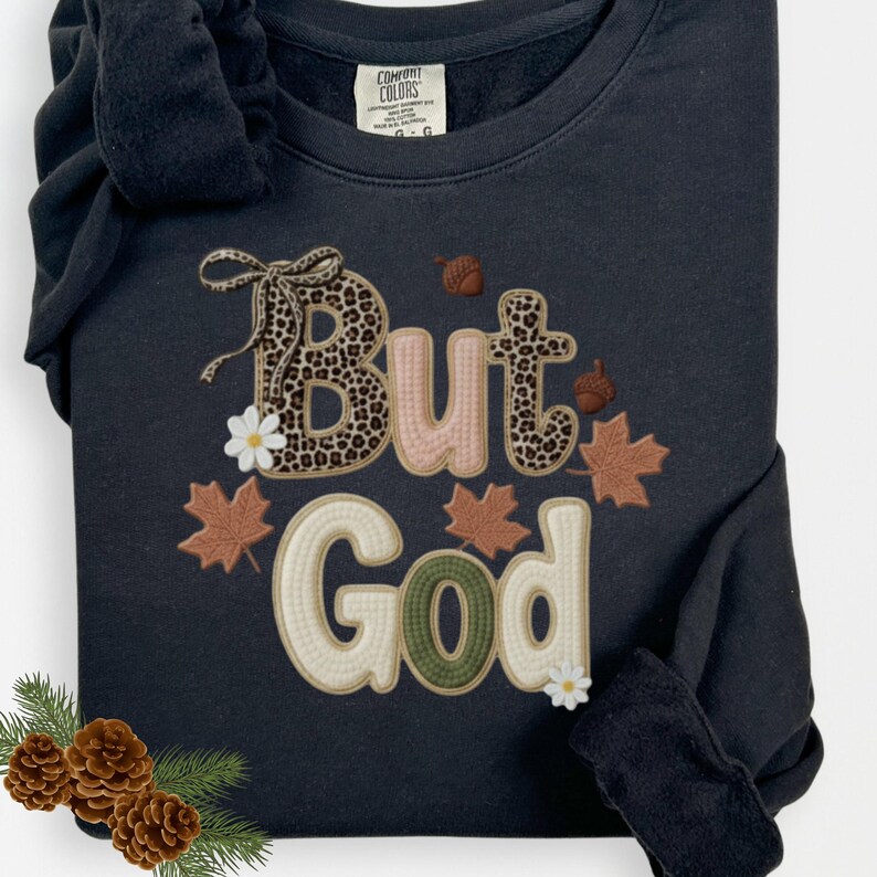 Pu&ograve; includere: Felpa nera con la frase "But God". La parola "But" presenta un motivo leopardato, mentre "God" &egrave; in verde e crema. Il design include elementi a tema autunnale come foglie e ghiande.