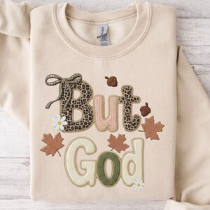 Pu&ograve; includere: Felpa beige con la frase ricamata "But God". La parola "But" presenta un motivo leopardato, mentre "God" &egrave; verde. Il design include elementi a tema autunnale come ghiande, foglie e margherite.