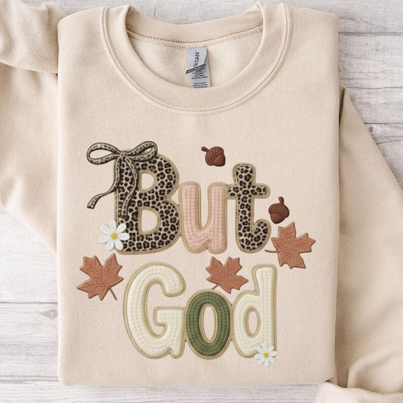 Pu&ograve; includere: Felpa beige con la frase ricamata "But God". La parola "But" presenta un motivo leopardato, mentre "God" &egrave; verde. Il design include elementi a tema autunnale come ghiande, foglie e margherite.