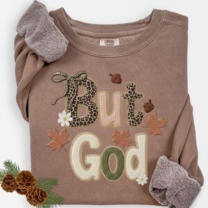 Pu&ograve; includere: Una felpa marrone con le parole "But God" in un carattere decorativo. La parola "But" ha un motivo leopardato e la parola "God" &egrave; in verde e bianco. La felpa presenta anche accenti a tema autunnale.