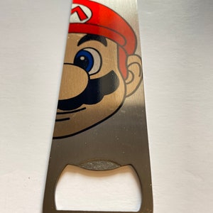 Puede incluir: Un abridor de botellas de plata con una imagen impresa de Mario de la serie de videojuegos Super Mario Bros. La cara de Mario es parcialmente visible, con su icónica gorra roja y ojos azules.
