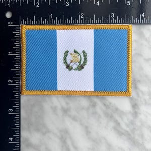 May include: Rectangular patch featuring the flag of Guatemala. The flag has vertical stripes of light blue and white, with the Guatemalan coat of arms in the center. The patch is bordered with gold stitching. The patch measures approximately 3 inches wide.