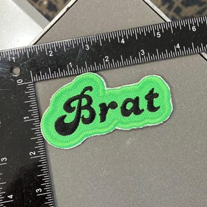 May include: Green and black embroidered patch with the word "Brat" in a cursive font.