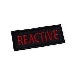 May include: Black rectangular patch with the word "REACTIVE" embroidered in red. The patch has a black border and is likely designed to be sewn or ironed onto clothing or accessories.