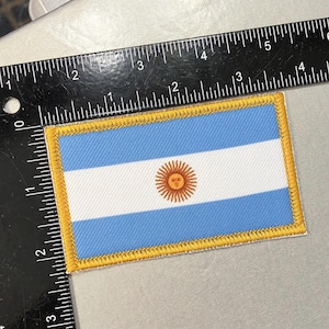 May include: A blue and white embroidered patch with a yellow border. The patch features the flag of Argentina with a sun in the center.