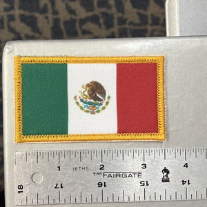 May include: A small embroidered patch of the Mexican flag with a yellow border. The flag is green, white, and red with the Mexican coat of arms in the center.