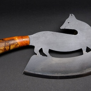 Può includere: Un grande coltello da macellaio curvo in metallo con una silhouette di volpe tagliata fuori dalla lama. Il manico è in legno con un motivo a marmo marrone e arancione.