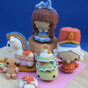 May include: A collection of six crocheted figures including a brown-haired doll with a blue bow, a white rocking horse, a gingerbread person, a tiered green Christmas tree, a soldier in a red hat, and a small white cake with a red flower.