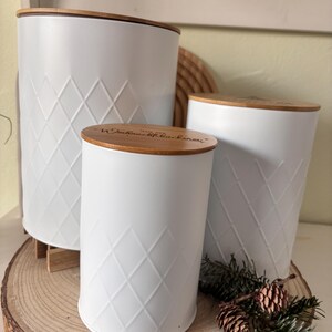 May include: Three white metal canisters with diamond pattern and wooden lids. The canisters are of varying sizes and are arranged on a wooden slice. The lids have text etched into them. The canisters are a decorative storage solution.