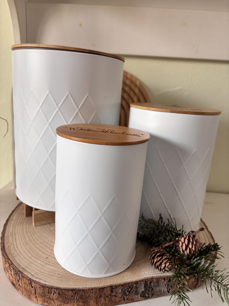 May include: Three white metal canisters with diamond pattern and wooden lids. The canisters are of varying sizes and are arranged on a wooden slice. The lids have text etched into them. The canisters are a decorative storage solution.
