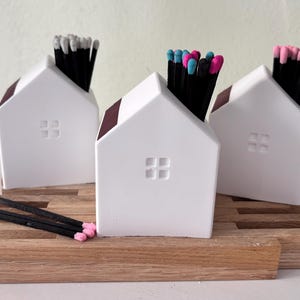 May include: Three white ceramic house-shaped match holders with maroon roofs and square window cutouts. Each holds a bundle of black matches with colored tips: white, pink, and a mix of pink and blue. The holders sit on a wooden base.