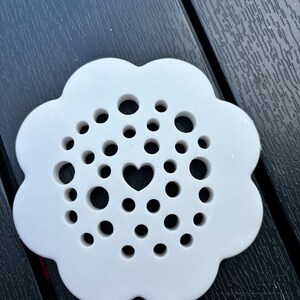 May include: White ceramic coaster with a flower-shaped design and a heart cutout in the centre. The coaster has multiple small holes around the heart.