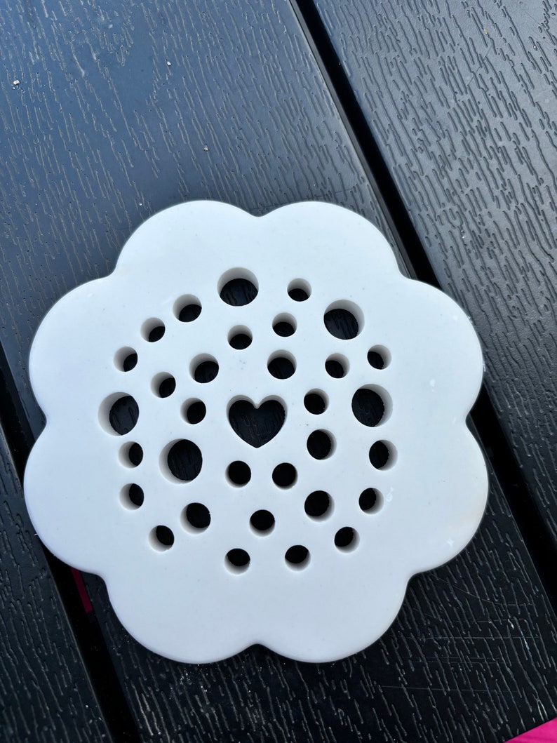 May include: White ceramic coaster with a flower-shaped design and a heart cutout in the centre. The coaster has multiple small holes around the heart.